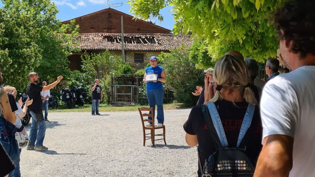 Ambiente esterno. Una donna è in piedi su una sedia e sorride con in mano una targa incorniciata, mentre un gruppo di persone intorno a lei sta applaudendo e scattando foto. Sullo sfondo si vedono edifici rurali storici.
