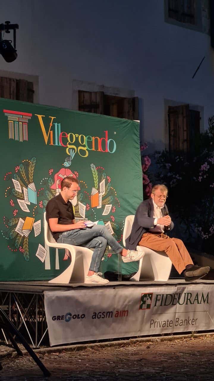 Due persone sedute su sedie bianche si trovano su un palco. Dietro di loro il fondale riporta il nome dell'evento "Villeggendo" e il logo. Foto scattata all'esterno che li inquadra di 3/4.